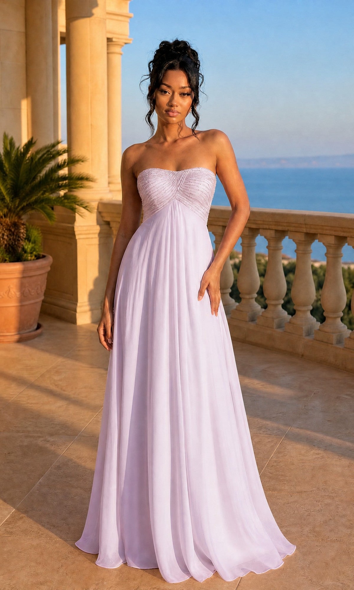 A woman in a strapless La Femme Dress 22363 in pale pink with an embellished bust stands on a sunny stone terrace with a balustrade, potted plant, and sea view in the background.
